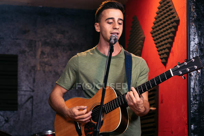 Jeune guitariste masculin positif jouant de la guitare acoustique et chantant au microphone dans un light club — Photo de stock