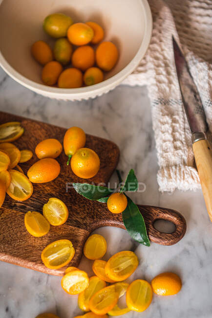 Montón vista superior de kumquats de corte naranja fresco sobre tabla de cortar de madera colocada sobre mesa de mármol con toalla en la cocina - foto de stock