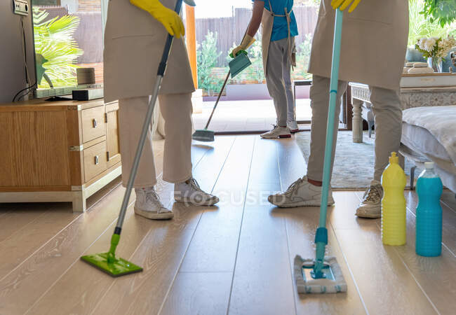 Cropped unrecognizable colleagues in aprons and gloves cleaning parquet floor with mops and brooms during work in modern spacious living room — Stock Photo
