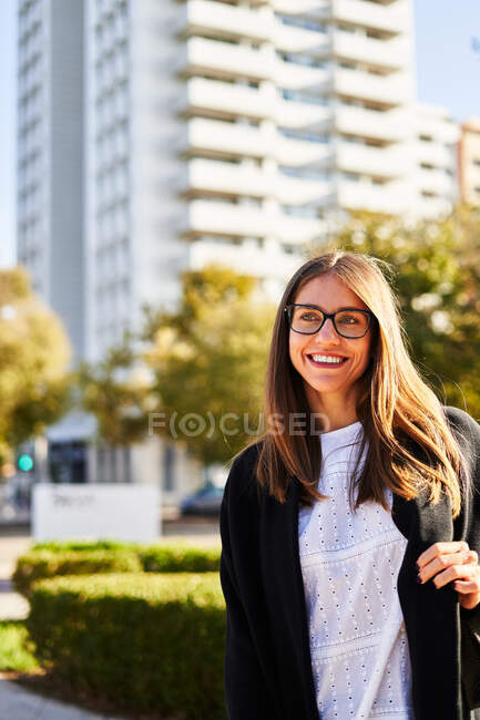 Mujer sonriente con cabello castaño en ropa casual y anteojos de pie en el parque con plantas verdes y mirando hacia otro lado contra los edificios en el distrito residencial de la ciudad en un día soleado - foto de stock