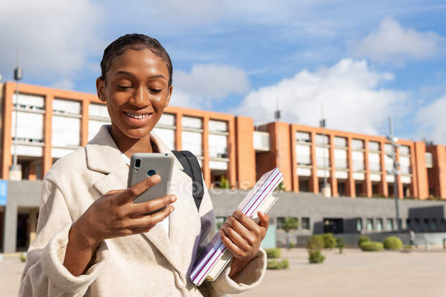 Studente afroamericana positiva con copybook messaggistica di testo sul cellulare mentre si trova nel campus universitario durante gli studi nella giornata di sole — Foto stock