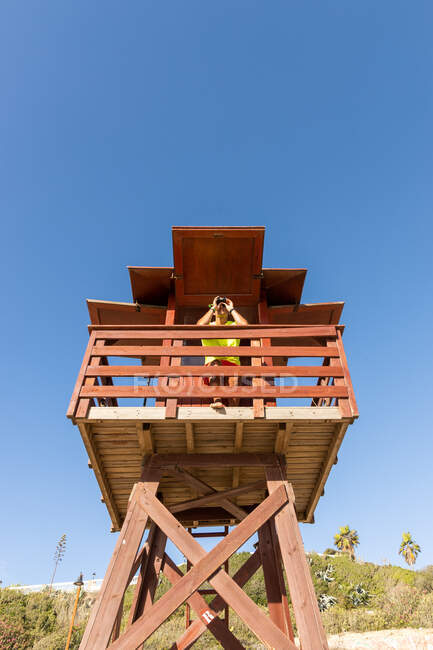 Basso angolo di guardare attraverso il binocolo sulla torre di guardia in legno mentre supervisiona la sicurezza in mare contro il cielo blu senza nuvole — Foto stock