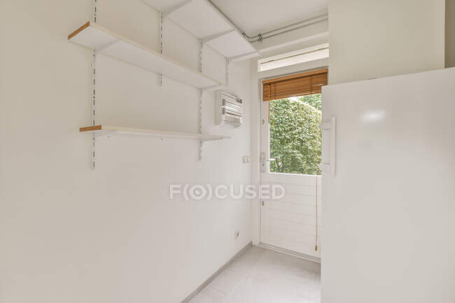 Enge Halle mit Regalen an weißer Wand gegen Tür mit Jalousie zu Hause bei Tageslicht — Stockfoto