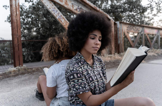 Concentrato afro-americano amiche seduti dietro e la lettura di libri sul vecchio ponte arrugginito — Foto stock