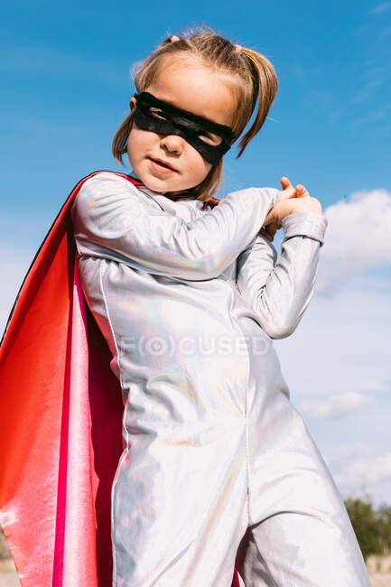 From below cute kid wearing masquerade superhero costume and eye mask standing against blue sky and looking at camera — Stock Photo