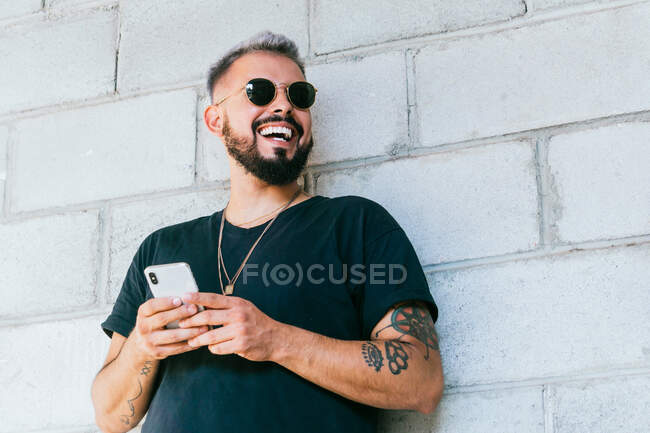 Счастливый бородатый парень с татуировками в черной футболке и солнцезащитных очках, стоящий у стены здания и использующий смартфон при дневном свете — стоковое фото