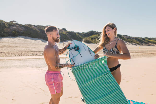 Sportive couple in swimwear taking off protective bag from surfboard while preparing for training on sandy beach in tropical resort — Stock Photo