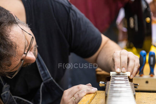 Обрезание мужского лютира в рабочей одежде и очках регулировка белого ореха на гитаре шея во время работы в профессиональной мастерской с оборудованием — стоковое фото