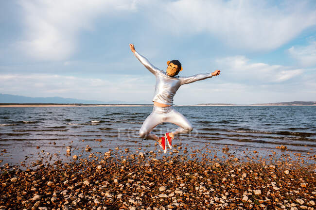 Unrecognizable male in monkey mask and silver clothes jumping with spread arms near wavy sea under cloudy blue sky — Stock Photo