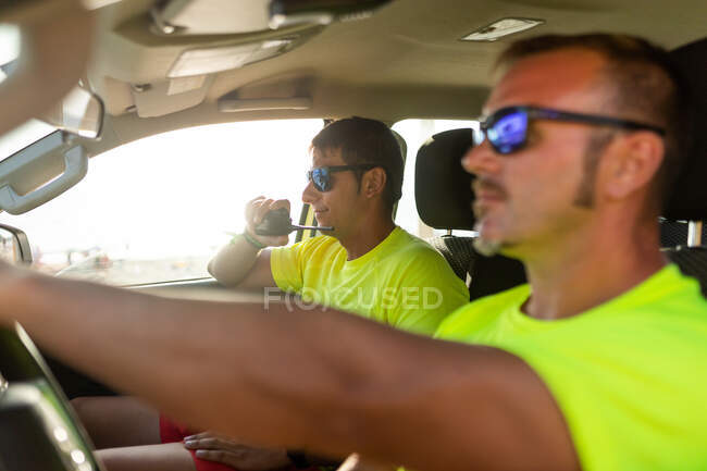 Vue latérale du maître nageur dans des lunettes de soleil conduisant une voiture près d'un homme parlant sur un émetteur-récepteur radio pendant la journée de travail — Photo de stock