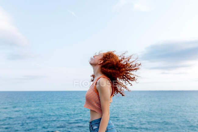 Side view of carefree anonymous female with eyes closed shaking curly ginger hair on coast of blue sea — Stock Photo