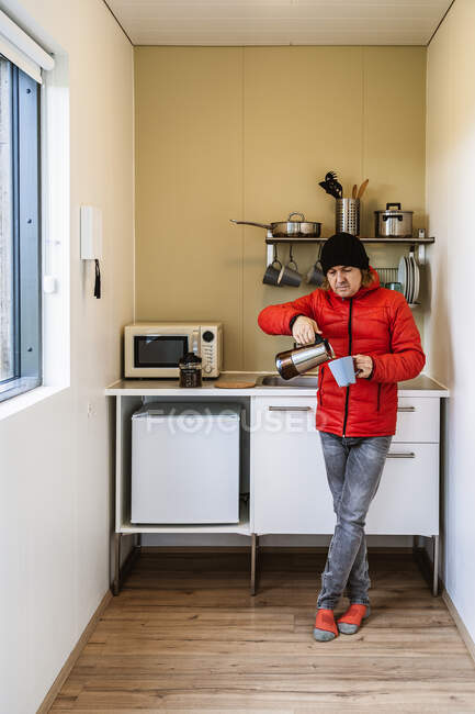 Повне тіло молодого чоловічого туриста в куртці і капелюсі, що наливає гарячу каву з термосу в чашку, стоячи на маленькій кухні — стокове фото