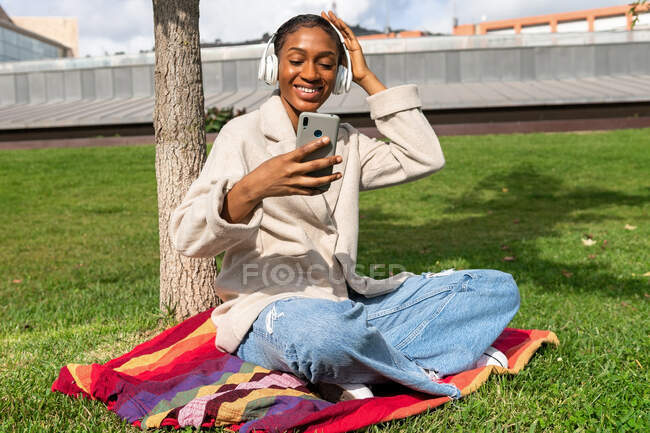 Corpo inteiro de conteúdo afro-americano feminino ouvindo música em fones de ouvido sem fio enquanto navega celular em xadrez perto do tronco da árvore no parque — Fotografia de Stock