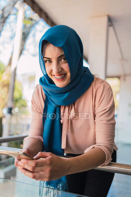 Entzückte Muslimin im Hidschab surft im Handy, während sie auf der Straße steht und in die Kamera schaut — Stockfoto