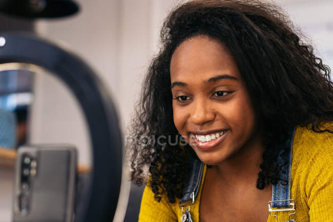 Lächelnde schwarze Frau auf der Couch, während sie ihr Smartphone an einer LED-Ringlampe in der Nähe professioneller Lichter auf Stativen nutzt — Stockfoto