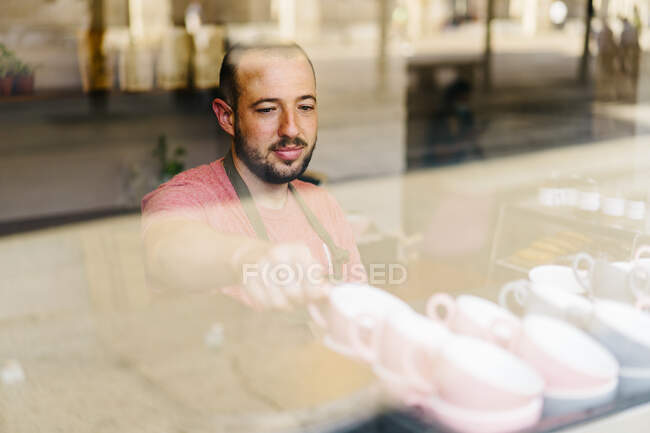 Através do vidro do trabalhador masculino no avental que toma copos da prateleira ao estar perto da janela durante o trabalho na cafetaria moderna — Fotografia de Stock