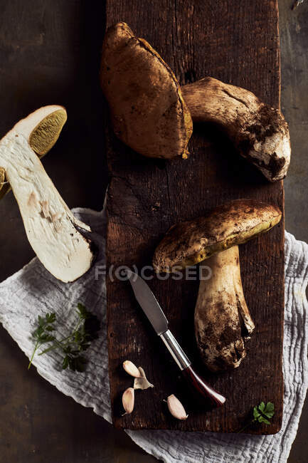 Vista superior de las setas Boletus edulis cortadas en bruto sobre tabla de cortar de madera con ajo y perejil en cocina ligera durante el proceso de cocción - foto de stock