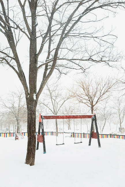 Parque infantil com balanços e slide coberto de neve entre árvores nuas no dia de inverno em clima sombrio em Madrid — Fotografia de Stock