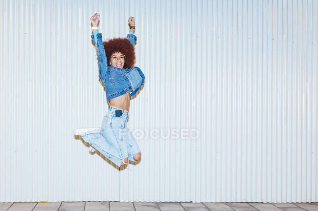 Full body of cheerful female with Afro hairstyle wearing stylish outfit jumping with raised arms on white background on street — Stock Photo