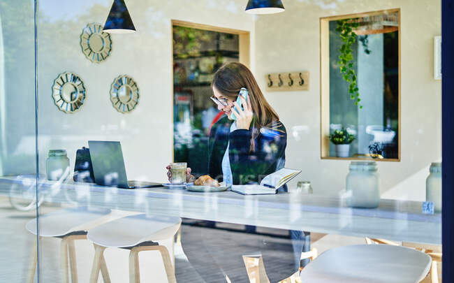 Attraverso vetro di donna d'affari che parla su smartphone a tavolo con caffè con cornetto vicino a computer portatile mentre lavora a distanza — Foto stock
