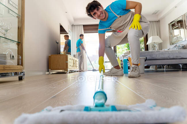 Ground level of cheerful young male professional cleaner in uniform and rubber gloves smiling while washing parquet floor with mop during work in spacious living room with colleagues — Stock Photo