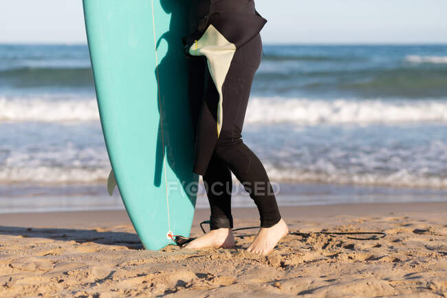 Side view of cropped unrecognizable female surfer in wetsuit with surfboard standing looking away on seashore washed by waving sea — Stock Photo