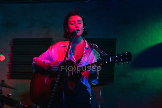 Confident lady with guitar with eyes closed singing in mic while performing song in bright club with neon light — Stock Photo