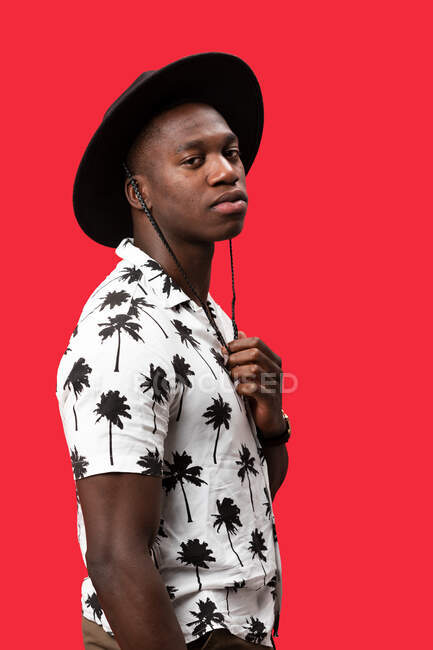 Side view of masculine self-confident African American male in stylish outfit standing against red background and looking at camera — Stock Photo