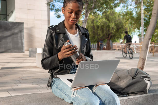 Contenido Mujer afroamericana freelancer en chaqueta de cuero abriendo termos con bebida caliente mientras está sentada con netbook en vueltas durante el trabajo en línea en la calle - foto de stock