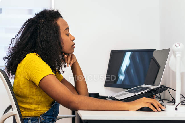 Vue latérale d'une femme afro-américaine assise à table et tapant sur un ordinateur portable dans une pièce lumineuse — Photo de stock