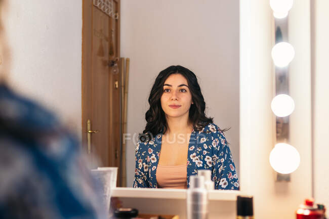 Jovem fêmea com penteado ondulado tocando o queixo enquanto se senta à mesa com produtos cosméticos e olhando para a reflexão no espelho no vestiário — Fotografia de Stock