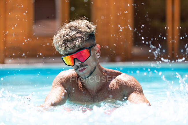 Homem sem camisa confiante com cabelo encaracolado em óculos de sol na piscina com água salpicante no dia ensolarado de verão no resort — Fotografia de Stock