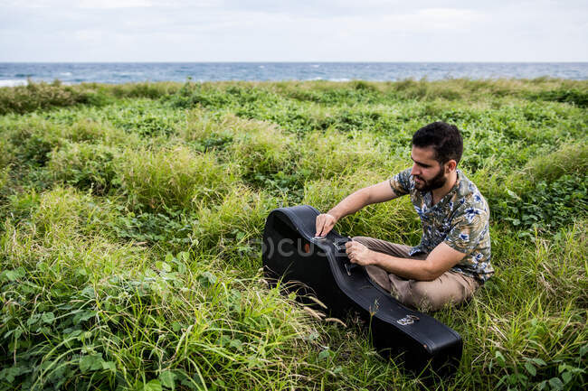 Сверху боковой вид спокойного мужчины-музыканта в повседневной одежде, сидящего на зеленой траве и открывающего черный ящик акустической гитары на берегу у моря при дневном свете — стоковое фото
