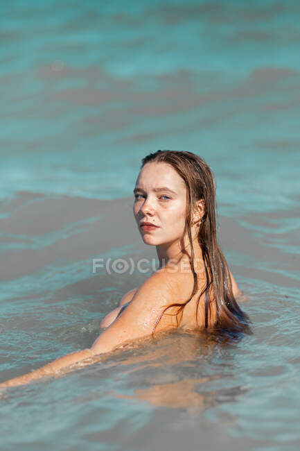 Mulher nua com cabelo molhado em pé na água do mar enquanto olha para a câmera sobre o ombro à luz do dia — Fotografia de Stock