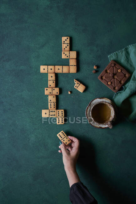 Top view of crop anonymous person eating sweet domino shaped cookies and drinking hot coffee on green background — Stock Photo