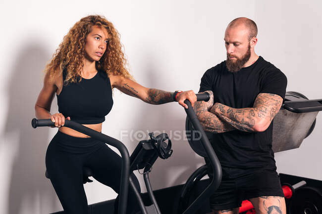 Sportive concentrée avec de longs cheveux bouclés assis sur une machine à vélo et faisant de l'entraînement cardio avec un entraîneur personnel dans la salle de gym — Photo de stock