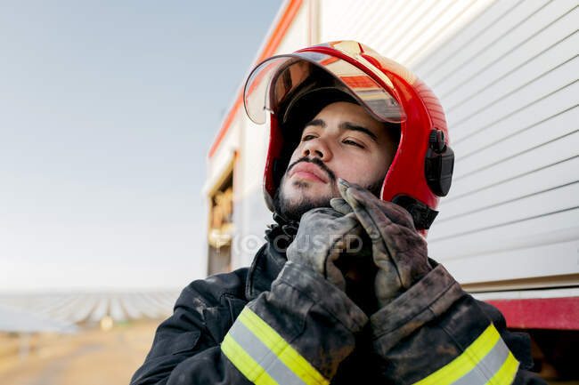 Jovem bombeiro barbudo vestindo uniforme com luvas de proteção olhando para longe enquanto regula a cinta do capacete em pé perto do carro de bombeiros no dia ensolarado — Fotografia de Stock
