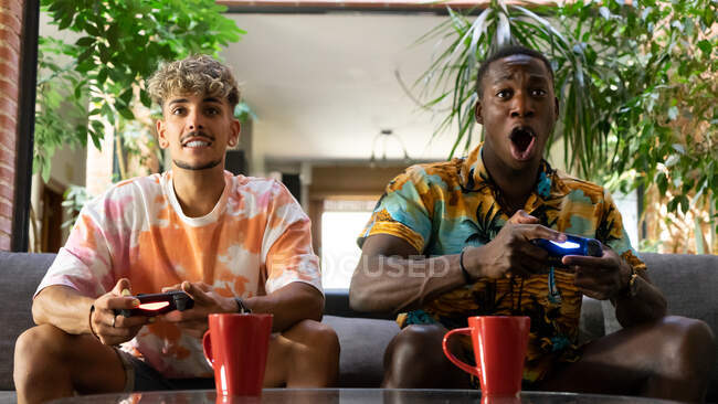 Diverse male friends with gamepads drinking beverages while sitting on couch and playing video game together in living room with green plants — Stock Photo