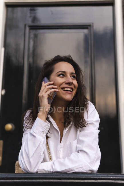 Vista lateral de uma jovem mulher feliz em roupas casuais de pé perto da entrada do edifício e apoiando-se no corrimão ao fazer um telefonema — Fotografia de Stock