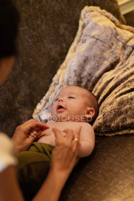 Dall'alto della madre senza volto del raccolto che si tiene per mano di adorabile bambino senza camicia sdraiato su un morbido cuscino sul divano di casa — Foto stock