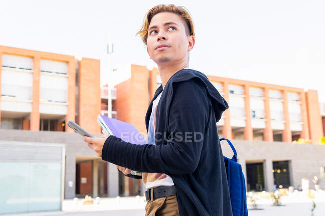 Cheerful male student in casual wear with copybooks text messaging on modern cellphone while standing near university building during studies — Stock Photo