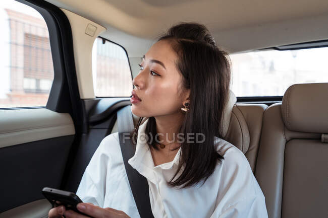 Азиатская пассажирка в формальной одежде просматривает мобильный телефон, сидя на заднем сиденье в такси и с интересом смотрит в сторону — стоковое фото