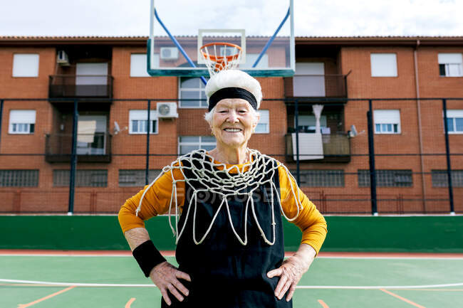 Sonriente hembra madura en ropa deportiva y red blanca mirando a la cámara mientras está de pie en el campo de deportes con aro de baloncesto durante el entrenamiento - foto de stock
