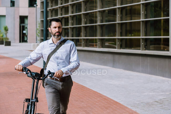 Исполнительный работник в формальной одежде, стоящий с велосипедом возле современного офисного здания — стоковое фото