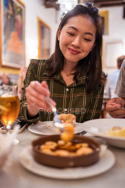 Conteúdo jovem fêmea étnica com longos cabelos escuros em roupas casuais sorrindo enquanto se senta à mesa no restaurante e come camarões deliciosos — Fotografia de Stock