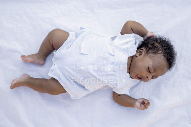 Сверху спокойный афроамериканец с темными вьющимися волосами в повседневной одежде, лежащий на белом одеянии и зевающий во сне днем — стоковое фото