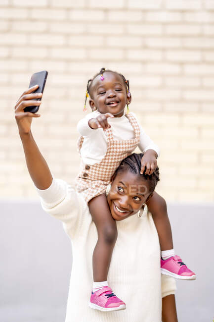 Веселая маленькая афроамериканка в стильном платье сидит на плечах улыбающейся матери, делая селфи на смартфоне на улице напротив кирпичной стены при дневном свете — стоковое фото