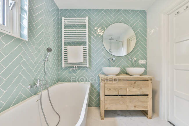 Banheira branca com mangueira colocada perto de pias na parede com espelho redondo na luz elegante banheiro com secador de toalhas e porta — Fotografia de Stock