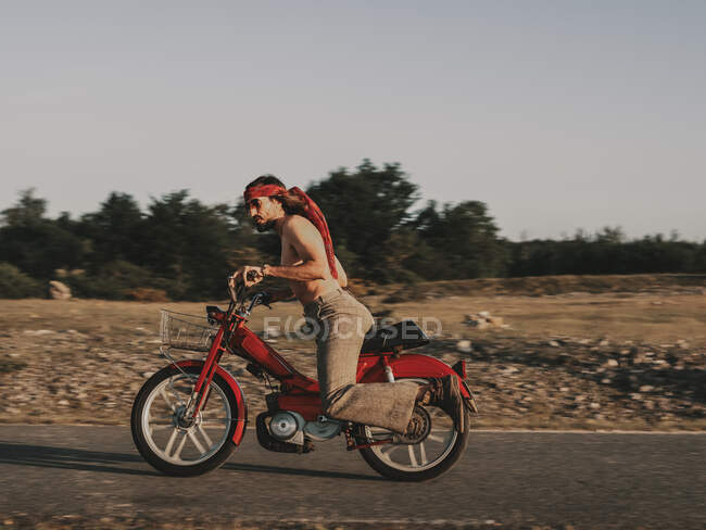 Full body side view of shirtless male hippie in headscarf riding moped on road during trip in nature on summer day — Stock Photo