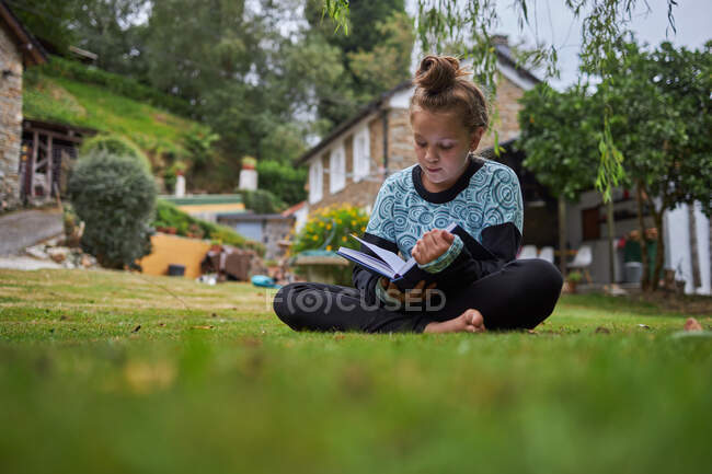 Cuerpo completo de chica descalza concentrada leyendo interesante libro mientras está sentado en el césped cubierto de hierba en el patio trasero contra el edificio residencial en el campo - foto de stock